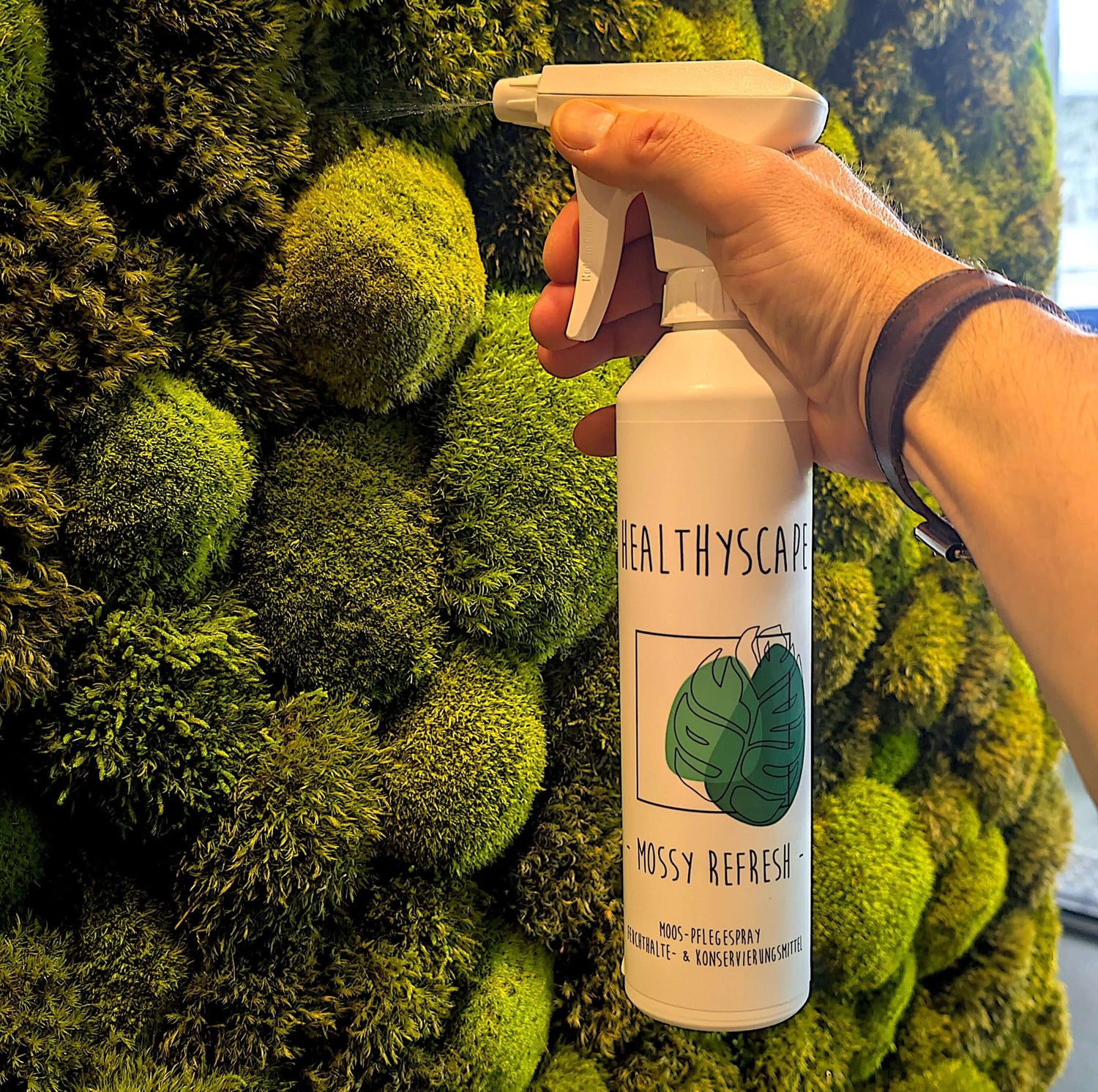 Hand holding a spray bottle labeled 'Mossy Refresh' in front of a vibrant green moss wall.