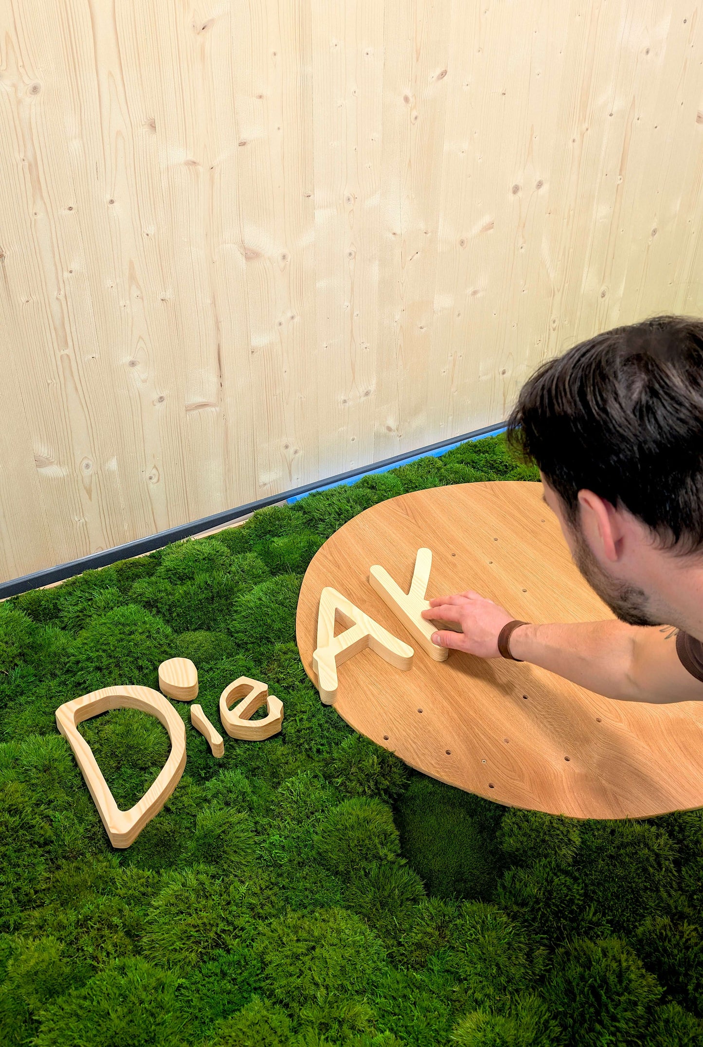 Ein Mann steckt einzelne Holzelemente eines Holzlogos auf ein Moosbild