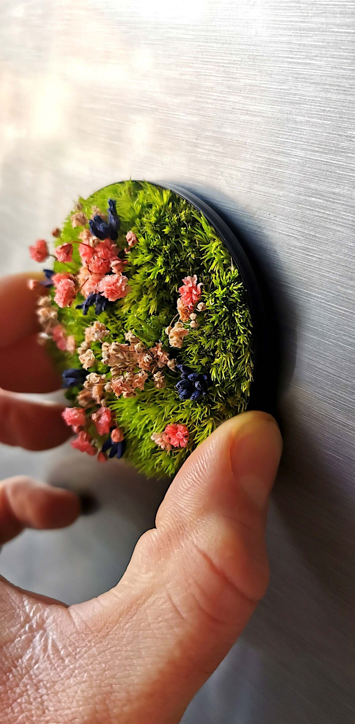 Hand placing a small round flower art piece titled 'Spring Vibes' on a metallic surface.