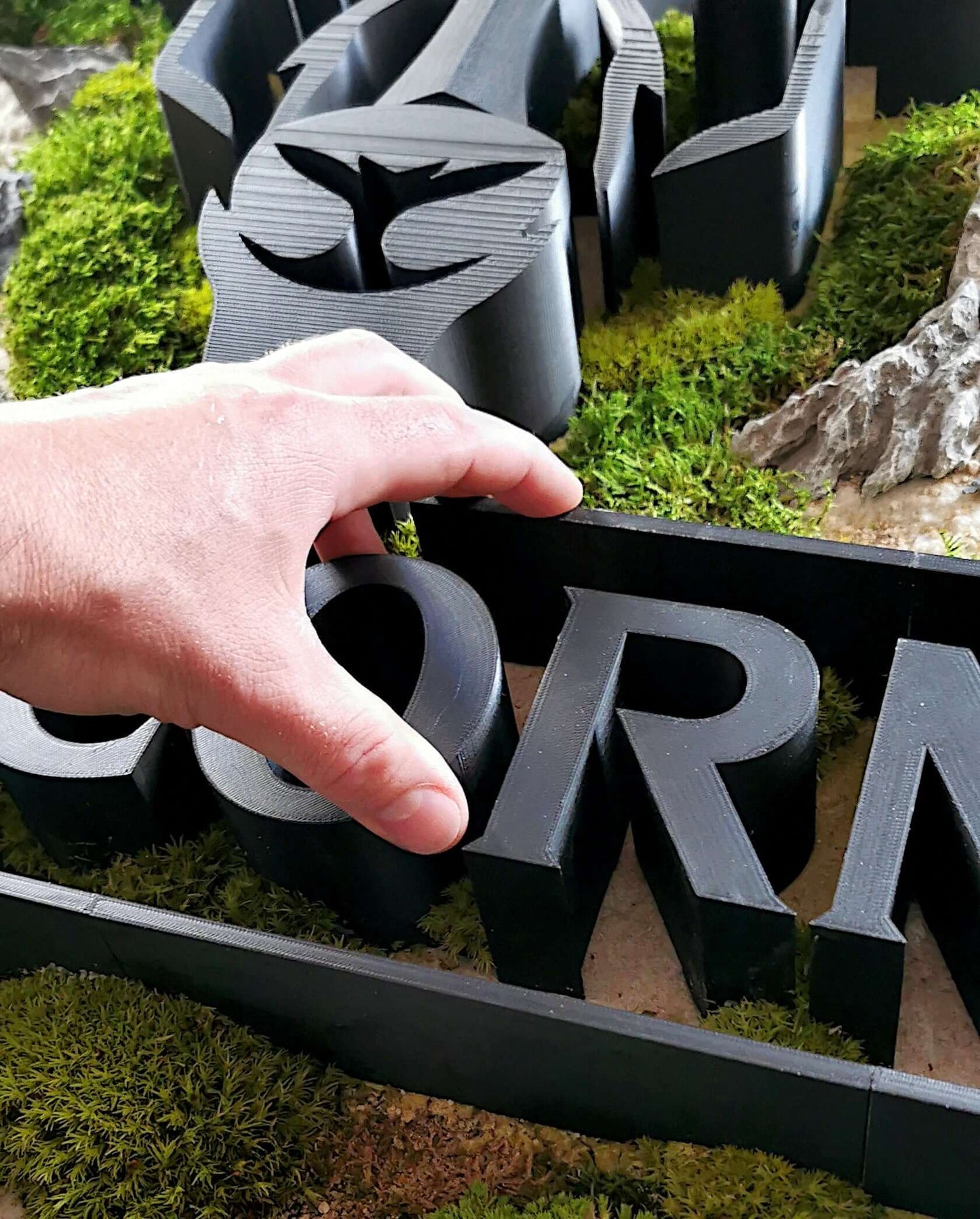 Moosbild mit Firmenlogo als 3D-Druck & Metallverblendung, Detailaufnahme eines Hands, das das 3D-Logo greift.