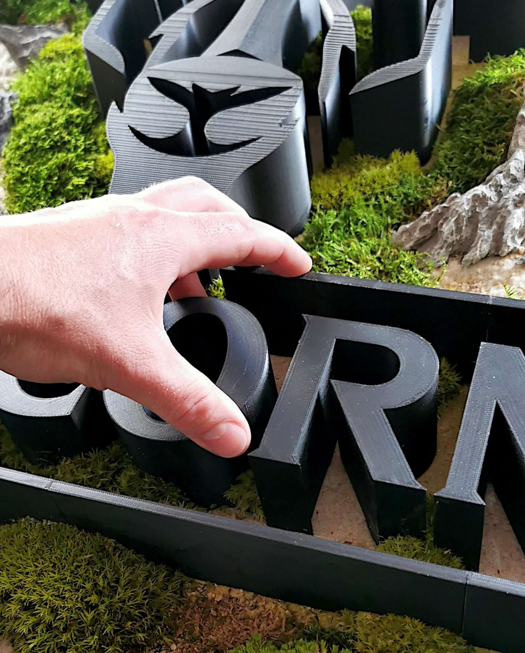 Moosbild mit Firmenlogo als 3D-Druck & Metallverblendung, Detailaufnahme eines Hands, das das 3D-Logo greift.